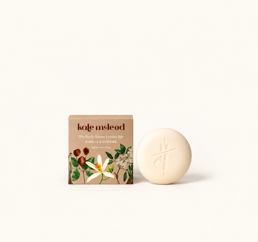 A round cream-colored body stone lotion bar with an embossed symbol, displayed next to its brown kraft paper packaging box featuring botanical illustrations, on a clean off-white background.