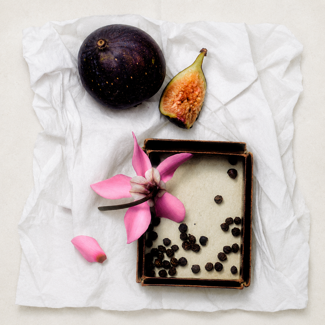 A styled flat-lay featuring a whole and halved purple fig, a pink flower, and small dark seeds arranged on a crumpled white paper sheet.