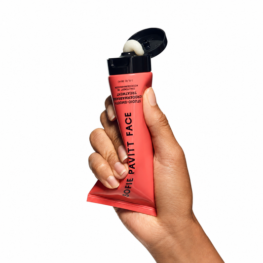 A hand holding a squeeze tube of Sofie Pavitt Face Lacquerise High Shine Acrylic Face Paint in a vibrant coral-red color, with the black cap open and showing a dollop of white paint, on a solid white background.