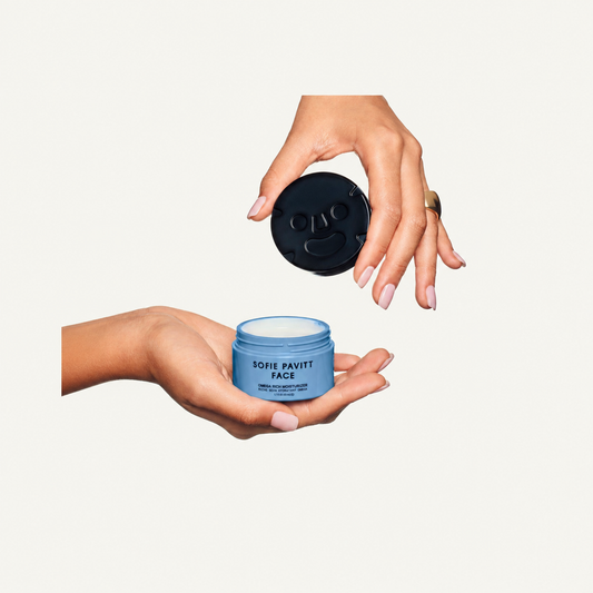 A person's hands holding a light blue jar labeled 'SOFT PAVITT FACE' with the black lid floating above, against a plain off-white background.