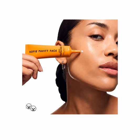 A close-up of a woman's face with an orange squeeze tube of Sofie Pavitt Face cream being applied to her cheek, set against a solid white background.