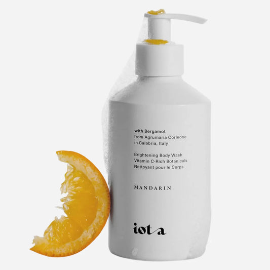 A white pump bottle of iot a Mandarin brightening body wash with a yellow-orange citrus slice next to it, on a light gray background.